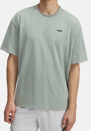 Person iført en lysegrøn oversized t-shirt med et lille sort mærke på brystet og en sølvkæde om halsen.