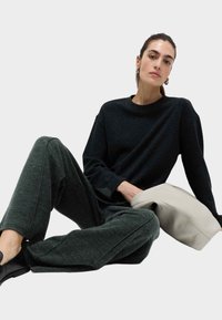 Czarna szarość długi rękaw top połączony z ciemnozielonymi spodniami w tapered. Miękkie faktury z subtelnymi wzorami. Trzyma jasnoszary woreczek. Czarne buty.