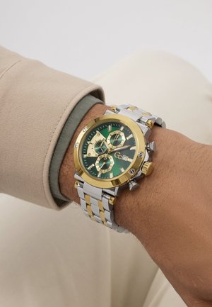 Käekell kuldse ja hõbedase metallrihmaga, roheline sihverplaat, kolm alamsihverplaati, kuupäeva näidik, kantud vasakul randmel beežide ja roheliste varrukatega.