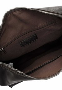 Intérieur d'un sac en cuir noir avec doublure en tissu brun, une poche zippée et une étiquette de marque. Détails de la fermeture éclair et des coutures visibles.
