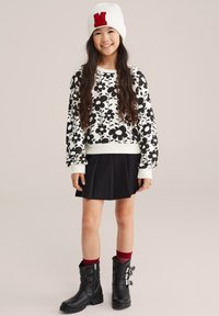 Mädchen mit langen Haaren, das einen weißen blumigen Pullover, einen schwarzen Rock, schwarze Stiefel, rote Socken und eine weiße Mütze mit rotem M trägt und lächelt.