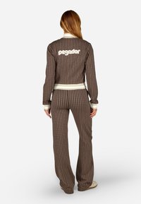 Brūna rakstaina treniņtērpa komplekts, kurā ir īsa jaka ar baltu logotipu un saskaņotas bikses ar plašām kājām un ribotām aprocēm.