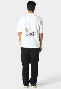 Valkoinen lyhythihainen T-paita, jossa on värikäs grafiikkaprintti, joka esittää palmun, auton ja raidoitetun taustan. Yhdistetty mustiin housuihin.