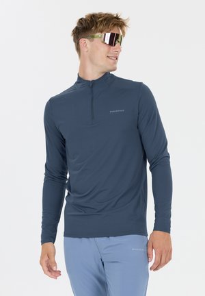 Man draagt een blauw sportief shirt met lange mouwen en lichtblauwe broek, kijkt zijwaarts, met reflecterende sportieve zonnebril.