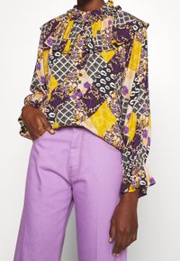 Blouse colorée avec un design en patchwork comprenant des motifs floraux et géométriques en violet, jaune et noir, un col froncé et des manches longues.