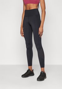 Høyt liv svarte leggings med en glatt tekstur, med perforerte detaljer nær anklene og sammen med svarte treningssko.