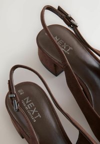 Rudos zomšos slingback aukštakulniai su blokuotu kulnu, su metaliniu sagtimi ir paminkštinta vidpadžiu. Prekės ženklas matomas ant vidpadžio.