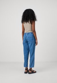 Kobieta z kręconymi włosami w beżowej bluzce bez rękawów, niebieskich spodniach z wysokim stanem i czarnych sandałach stojąca na jednolitym tle.