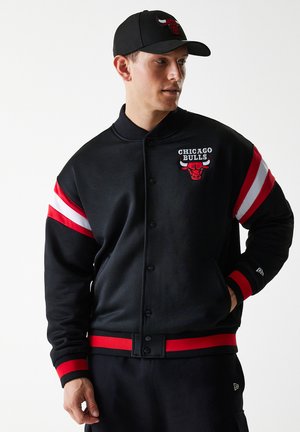 Juodos spalvos Čikagos "Bulls" universiteto striukė, turinti raudonus ir baltus dryžuotus rankovėmis, siuvinėtą logotipą ir gumuotus rankogalius bei apačią. Matinis paviršius.