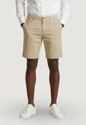 Hombre con pantalones cortos beige hasta la rodilla, una camisa blanca de manga larga y zapatillas blancas, de pie con las manos en los bolsillos.