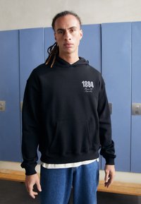 Hoodie noir en coton, avec une poche avant, des poignets côtelés et un imprimé comprenant « 1894 » en blanc sur le coin supérieur gauche.