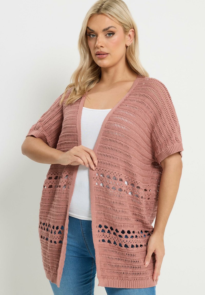 Frau mit blondem Haar, die eine staubrosafarbene, offene Strickjacke über einem weißen Oberteil und blauen Jeans trägt, steht vor einem einfarbigen hellen Hintergrund.