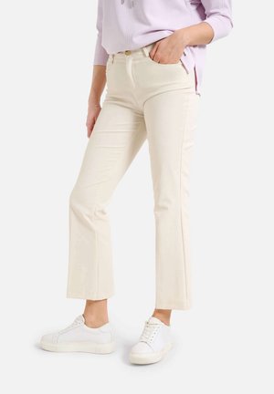 Persona con pantalones rectos color crema, zapatillas blancas y una camiseta de manga larga color lavanda claro, con una mano en el bolsillo.