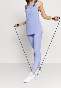 Světle fialový atletický top bez rukávů a legíny s vzorovaným ramínkem, doplněné bílými teniskami a drženým černým švihadlem.