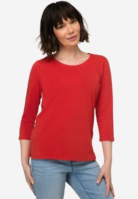 Camicia rossa a maniche lunghe con scollatura tonda e spacchi laterali, realizzata in morbido tessuto di cotone. Abbinata a jeans azzurri chiari. Design semplice e casual.