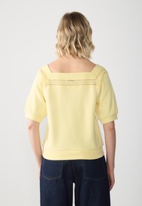 En blond kvinna vänd bort med en ljusgul top med fyrkantig halsringning och ögla spetsdetalj, samt mörkblå jeans mot en vit bakgrund.