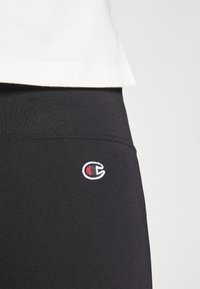 Svarta stretchleggings med en fittad design, prydda med en liten broderad logotyp i vitt och rött på höften. Slät tygtextur.