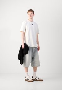 T-shirt bianca in cotone con un logo quadrato, pantaloni shorts in denim blu chiaro, maglione nero appoggiato, scarpe bianche e marroni con strisce nere.