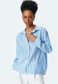 Camisa de botón azul claro con rayas verticales en un azul más oscuro y negro. Presenta un cuello, mangas largas y un corte holgado.