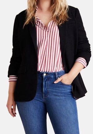 Frau trägt schwarzen Blazer über einem rot-weiß gestreiften Hemd, das in blaue Jeans gesteckt ist, mit einer Hand in der Tasche und der anderen entspannt.