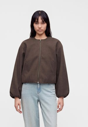 Frau trägt eine dunkelbraune Reißverschlussjacke mit langen, bauschigen Ärmeln und hellblauen Jeans, steht vor einem einfarbigen grauen Hintergrund.