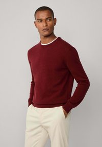 Muškarac u bordo puloveru s okruglim izrezom preko bijele košulje i bež hlačama, stoji s rukama u džepovima naslonjen na jednobojnu pozadinu.