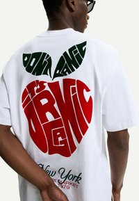 T-shirt bianco in cotone con testo grafico rosso e verde. Il design include una forma a cuore e la scritta "NON PANICARE", con "è ORG NIC" sotto.