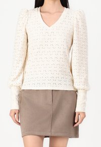 Kvinna som bär en krämfärgad stickad långärmad V-ringad tröja med puffärmar och en taupe över-knä kjol.