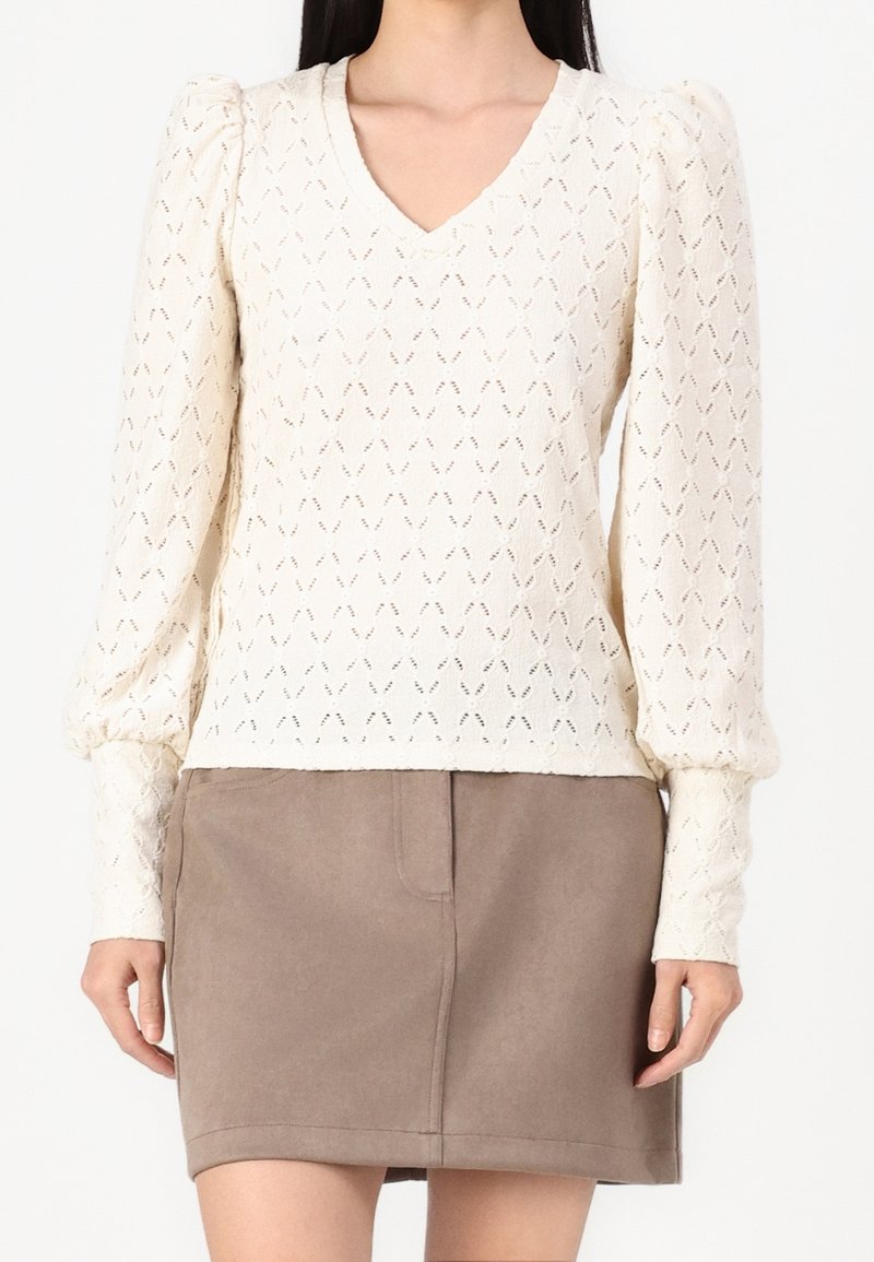 Kvinna som bär en krämfärgad stickad långärmad V-ringad tröja med puffärmar och en taupe över-knä kjol.