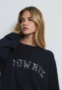 Pull marin à la coupe décontractée, présentant un design en sequins "BROWNIE" sur la poitrine. Texture en tricot et manches larges.