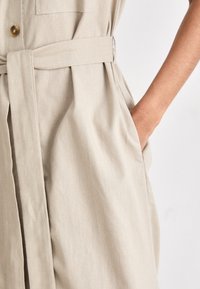 Μπεζ λινό φόρεμα με κουμπιά μπροστά, ζώνη στη μέση και πλαϊνές τσέπες, με χαλαρή και ρέουσα γραμμή.