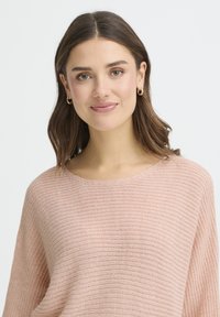 Pull tricoté rose clair avec une coupe ample et un large décolleté. Texture lisse avec de fines rayures horizontales. La femme porte de petites boucles d'oreilles créoles.