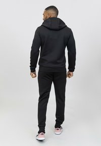 Un sweat à capuche noir et un jogging, fabriqués en tissu lisse. Le sweat à capuche est doté d'un cordon de serrage et de poignets côtelés. Les chaussures sont blanches avec des accents roses.
