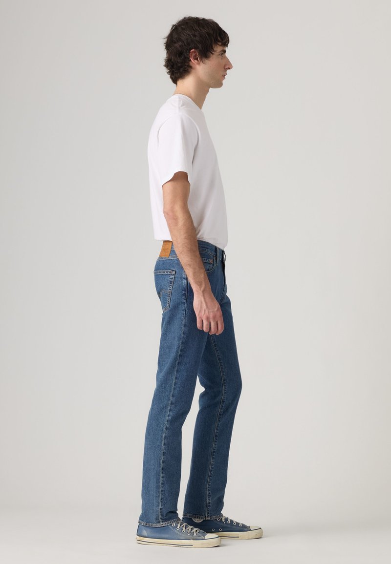 Hombre de pie de lado con camiseta blanca, vaqueros azules y zapatillas azules sobre un fondo liso gris claro.