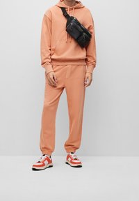 Pfirsichfarbene Kapuzenjacke und Jogginghose aus einem weichen Stoff, mit gerippten Bündchen und einer schwarzen Umhängetasche mit Reißverschlussfächern. Rote und weiße Sneakers.