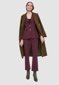 Abito color mogano con giacca su misura e pantaloni abbinati, indossati sotto un lungo cappotto verde oliva, abbinati a stivaletti verde oliva.