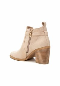 Botín de ante beige a la altura del tobillo con punta redonda, cremallera lateral y tacón de madera apilada. Cuenta con una asa en la parte posterior.