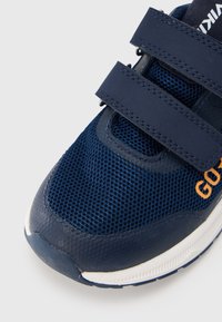 Αθλητικό παπούτσι σε ναυτικό μπλε με επάνω μέρος από δίχτυ, διπλές λουριά Velcro, καουτσούκ σόλα και πορτοκαλί λογότυπο. Στρογγυλεμένος σχεδιασμός μύτης.