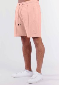 Pfirsichfarbene Shorts aus weichem Stoff, mit einem Kordelzugbund, Seitentaschen und einem Logo, das nahe dem Saum bedruckt ist.