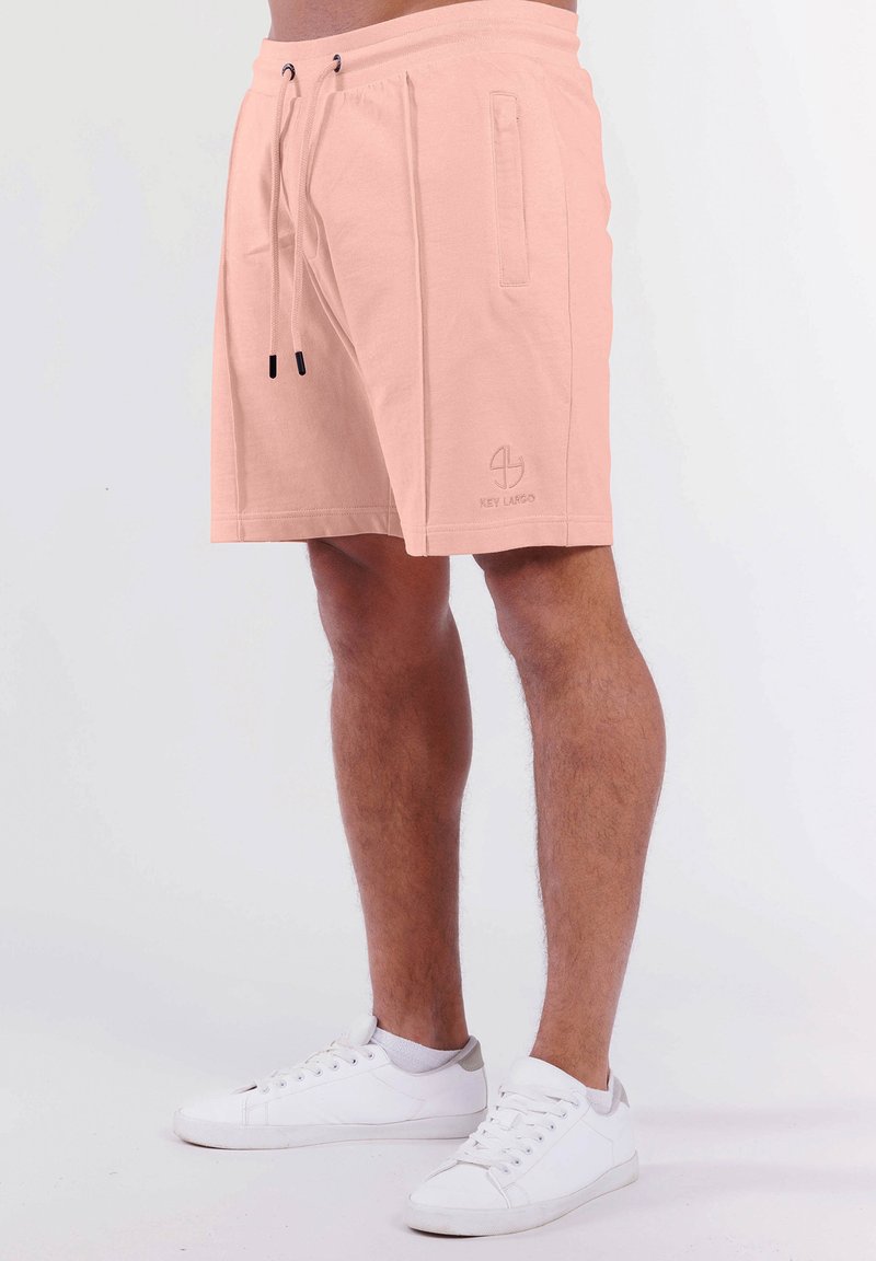Pfirsichfarbene Shorts aus weichem Stoff, mit einem Kordelzugbund, Seitentaschen und einem Logo, das nahe dem Saum bedruckt ist.