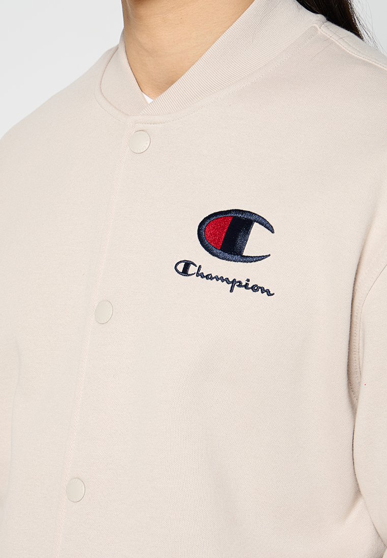 Giacca varsity beige Champion con bottoni a pressione, colletto a costine e logo ricamato in navy, rosso e nero. Materiale in cotone liscio.