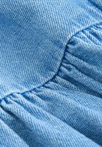 Ljusblå denimtyg med en strukturerad väv, med inslag av rynkade sektioner och synliga sömnadsdetaljer. Mjuk textur med en avslappnad finish.