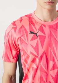 Camiseta deportiva de manga corta en rosa brillante con un patrón angular y acentos en negro. Presenta el logo de Puma en el frente y un panel lateral negro en contraste.