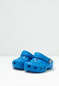 Pár modrých nazouvacích dřeváků s ventilačními otvory a nastavovacími patními pásky na bílém povrchu s jednobarevným pozadím.