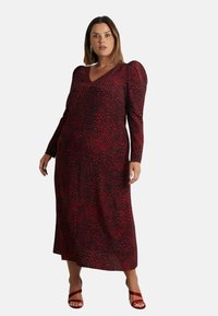 Dlouhé šaty s černo-červeným leopardím vzorem a výstřihem do V, které mají přiléhavou siluetu a nabírané ramena, doplněné červenými sandály.