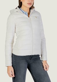 Giacca trapuntata bianca con zip frontale, cappuccio e dettaglio con logo; presenta una vestibilità aderente e una texture liscia. Abbinata a jeans scuri.