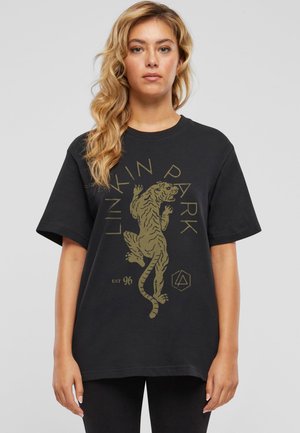 Frau mit langen blonden Haaren trägt ein schwarzes Linkin Park T-Shirt mit einer Grafik eines kletternden Tigers und Text auf einem einfarbigen grauen Hintergrund.
