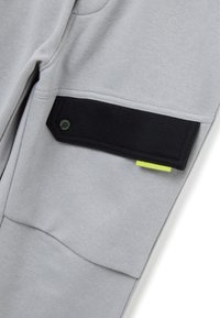 Pantalones de chándal grises con un bolsillo de velcro negro con un logotipo de acento verde y una etiqueta neón amarilla, hechos de una tela suave y texturizada.