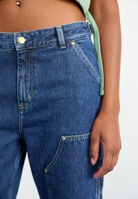 Primer plano de la cintura y la mano de una persona que lleva jeans de mezclilla azules con remaches dorados y una parte visible de una blusa verde.