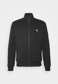 Giacca nera con zip e colletto alto, tasche laterali, polsini a coste e logo CK bianco sul lato sinistro del petto. Tessuto dalla consistenza liscia.